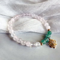 I Am Love” heart chakra bracelet by Moksha Jewelry, handcrafted with rose quartz and green aventurine beads, accented with gold details and a lotus charm, styled on a soft gray surface with a textured white fabric—designed to inspire love, balance, and emotional healing.