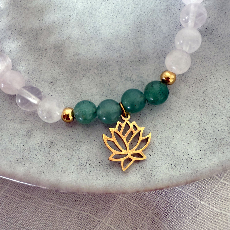 Close-up of the “I Am Love” bracelet by Moksha Jewelry, featuring green aventurine and rose quartz beads with gold accents and a lotus charm, displayed on a soft stone plate and sheer fabric—designed to open the heart chakra and promote love and healing energy.