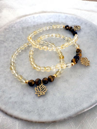 Set of “I Am Strong” chakra bracelets handcrafted by Moksha Jewelry, featuring golden citrine and tiger eye beads with gold-plated lotus charms, displayed on a light stone plate—designed to support personal power, clarity, and Manipura chakra energy.