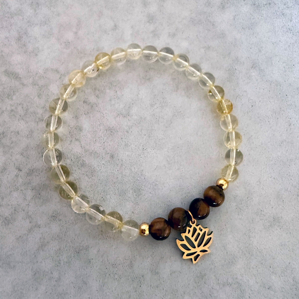 Moksha Jewelry’s “I Am Strong” chakra bracelet featuring citrine and tiger eye beads with a gold lotus charm, displayed on a soft stone surface—designed to support confidence, clarity, and empowerment through the Manipura chakra.
