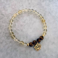Moksha Jewelry’s “I Am Strong” chakra bracelet featuring citrine and tiger eye beads with a gold lotus charm, displayed on a soft stone surface—designed to support confidence, clarity, and empowerment through the Manipura chakra.