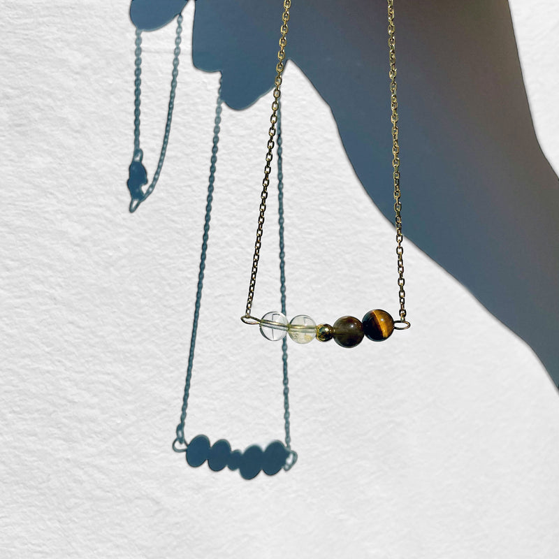 A fine golden necklace with 2 citrine beads, 2 tiger eye beads and one golden bead in the middle in front of a neutral background.