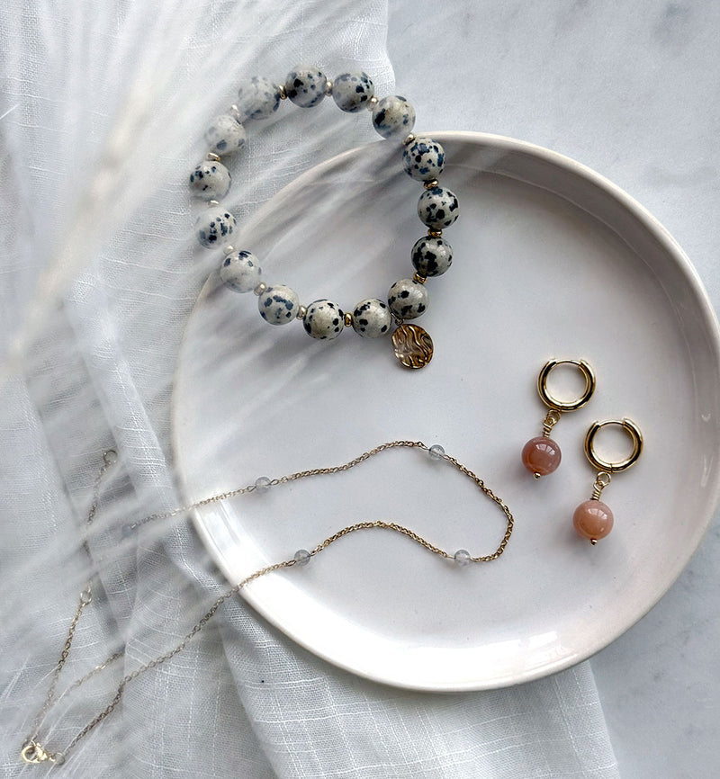 Flat lay of handcrafted gemstone jewelry on a ceramic plate, featuring a Dalmatian Jasper bracelet, peach moonstone hoop earrings, and a delicate moonstone necklace with gold accents on soft white fabric.