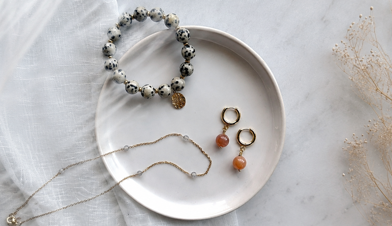 Flat lay of handcrafted gemstone jewelry on a ceramic plate, featuring a Dalmatian Jasper bracelet, peach moonstone hoop earrings, and a delicate moonstone necklace with gold accents on soft white fabric.