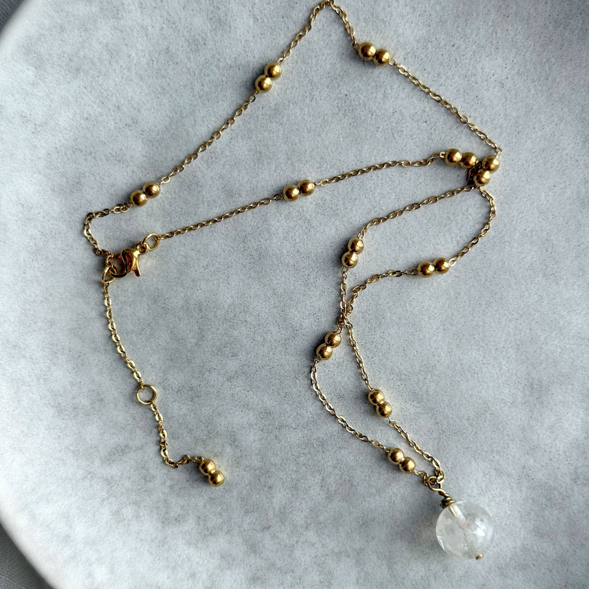 Gold-plated beaded chain necklace with a clear quartz pendant laid flat in a loose arrangement on a grey ceramic plate.