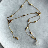 Gold-plated beaded chain necklace with a clear quartz pendant laid flat in a loose arrangement on a grey ceramic plate.