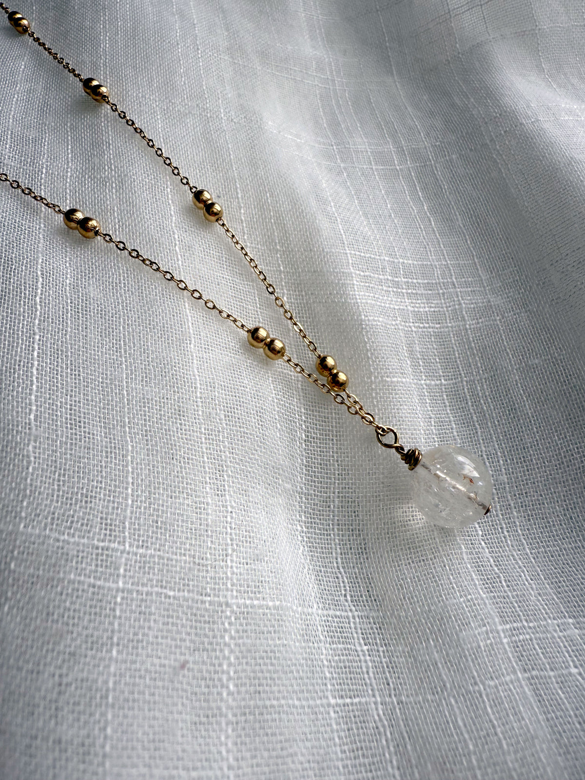 Close-up of the Lumina Necklace in clear quartz, showcasing a single round gemstone bead with natural inclusions, suspended from a fine gold-plated chain adorned with delicate gold balls, displayed on a soft white textured fabric.