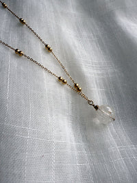 Close-up of the Lumina Necklace in clear quartz, showcasing a single round gemstone bead with natural inclusions, suspended from a fine gold-plated chain adorned with delicate gold balls, displayed on a soft white textured fabric.