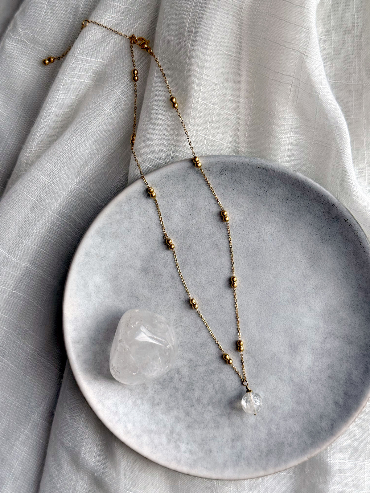 Full view of a gold-plated beaded chain necklace with a clear quartz pendant, styled on a grey ceramic plate with a polished clear quartz stone, set against a white textured fabric background.