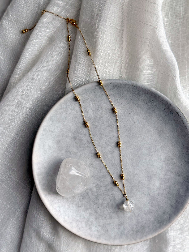 Full view of a gold-plated beaded chain necklace with a clear quartz pendant, styled on a grey ceramic plate with a polished clear quartz stone, set against a white textured fabric background.