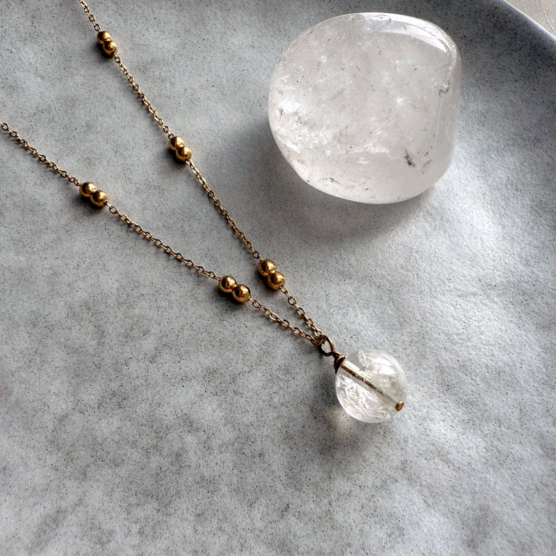 Close-up of a gold-plated beaded chain necklace with a round clear quartz pendant, displayed on a soft grey ceramic plate next to a polished clear quartz stone.