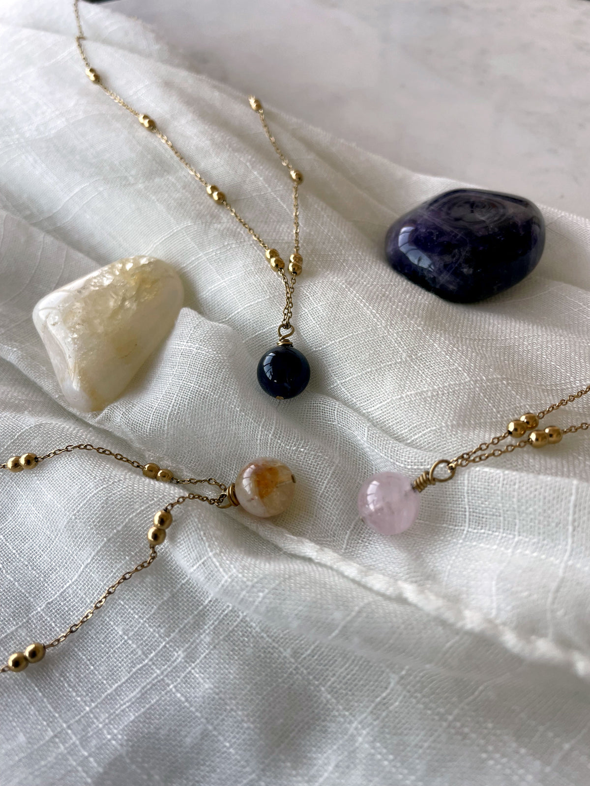 3 necklaces, with a 10mm gemstone bead, on made of amethyst, one made of citrine and one made of rose quartz on a gold plated necklace on a light linen fabric.