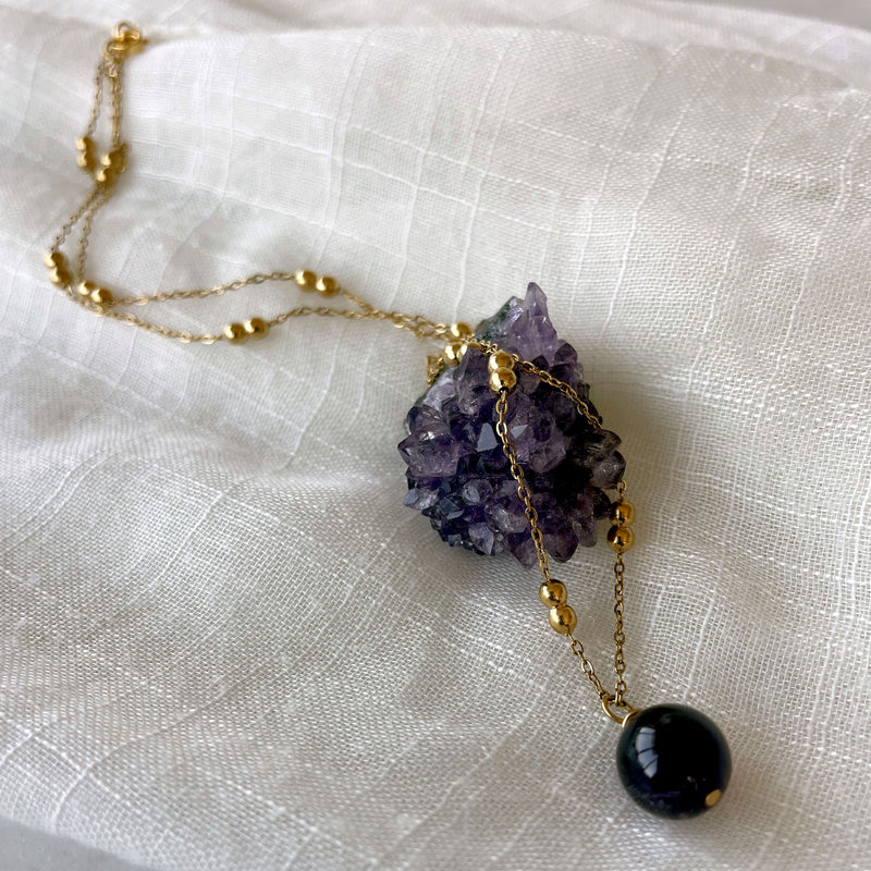 A close-up of gold plated necklace with a 10mm Amethyst bead on a delicate chain with 3mm Ball and 1mm Curb Chain on a light linen fabric on an amethyst druse.
