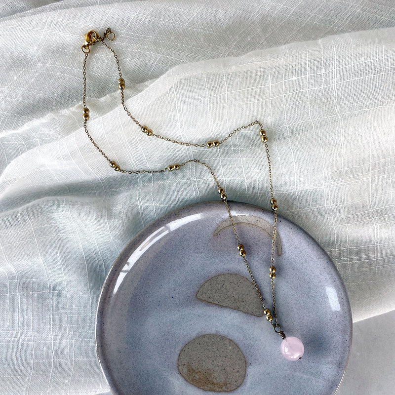 A gold plated necklace with a 10mm Rose Quartz bead on a delicate chain with 3mm Ball and 1mm Curb Chain on a ceramic plate.