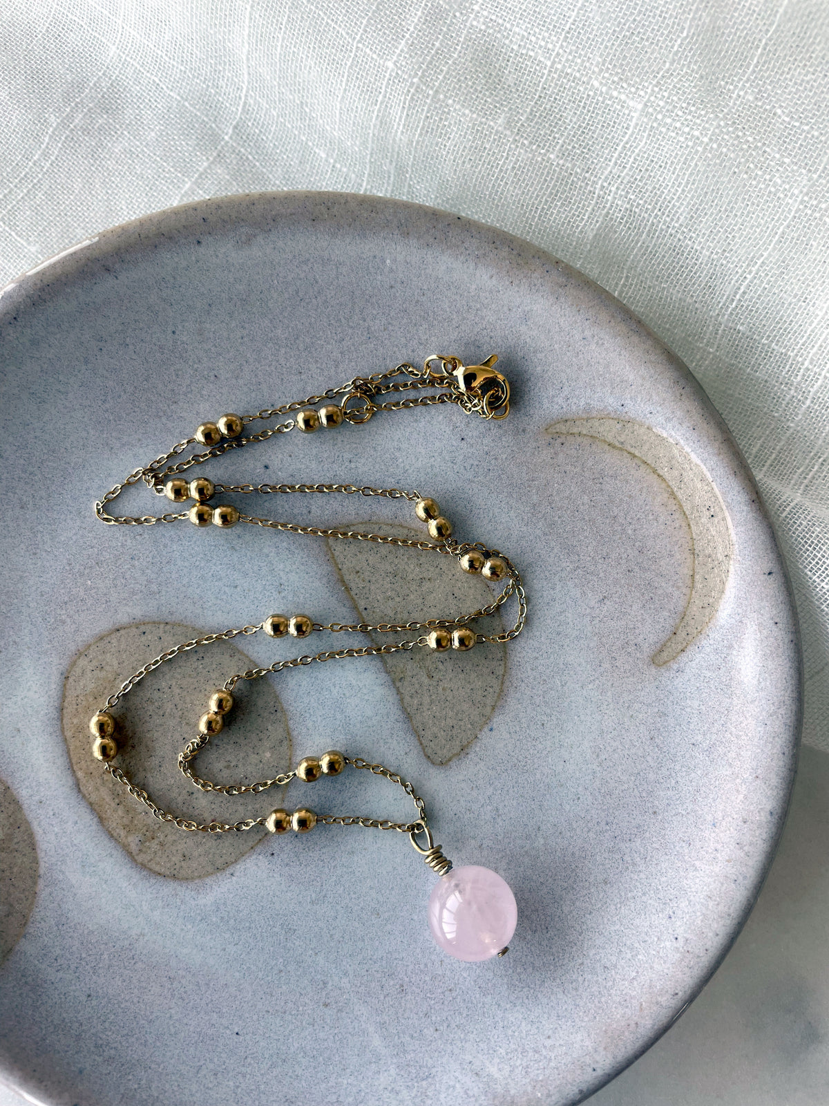 A gold plated necklace with a 10mm Rose Quartz bead on a delicate chain with 3mm Ball and 1mm Curb Chain on a ceramic plate.