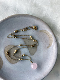 A gold plated necklace with a 10mm Rose Quartz bead on a delicate chain with 3mm Ball and 1mm Curb Chain on a ceramic plate.