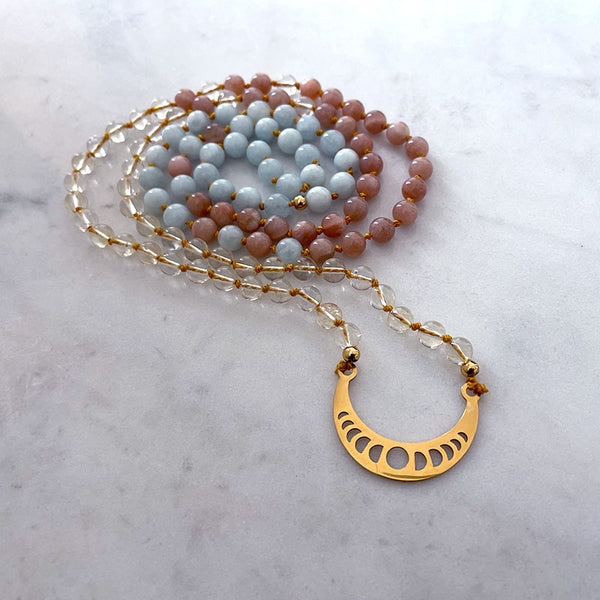 A mala made of citrine, peach moonstone and aquamarine beads with a gold-plated pendant in the shape of a crescent moon with the phases of the moon punched out.