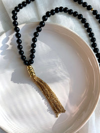 A close-up of a Mala made of 6mm black Tourmaline beads with a gold plated stainless steel chain tassel on a white ceramic plate.