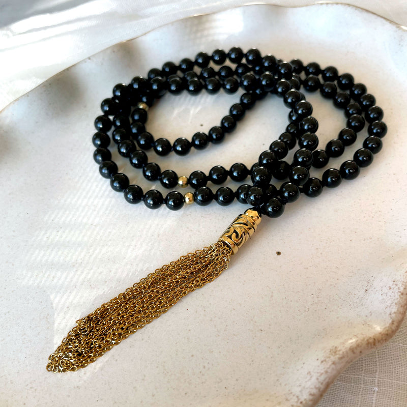A close-up of a Mala made of 6mm black Tourmaline beads with a gold plated stainless steel chain tassel on a gray ceramic plate.