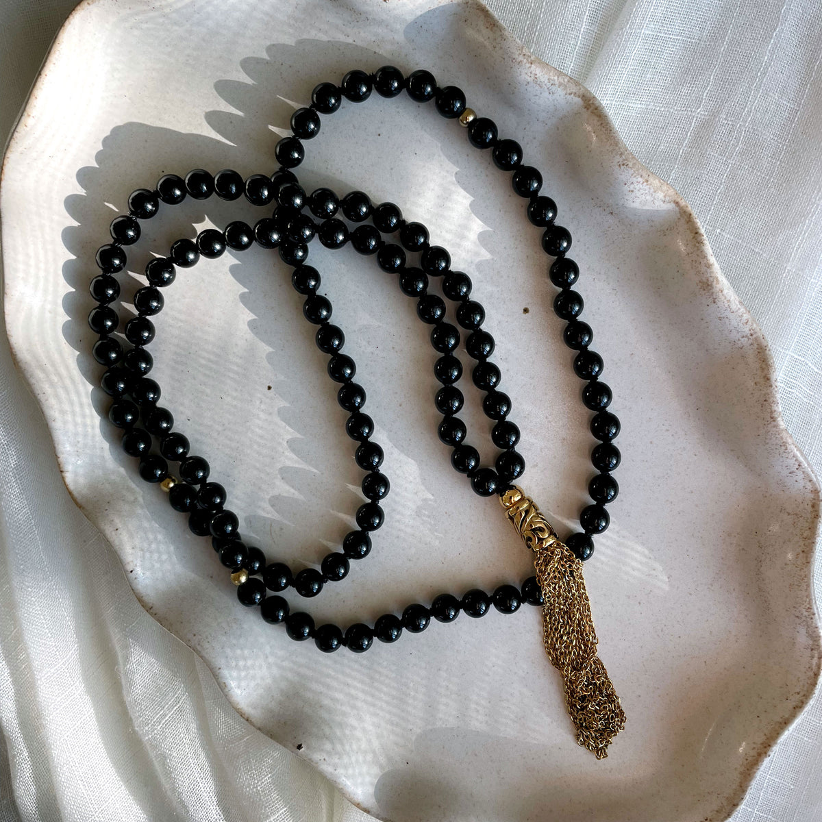 A Mala made of 6mm black Tourmaline beads with a gold plated stainless steel chain tassel on a gray ceramic plate.