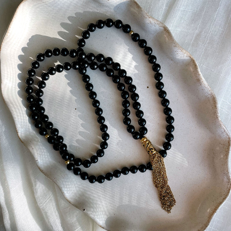 A Mala made of 6mm black Tourmaline beads with a gold plated stainless steel chain tassel on a gray ceramic plate.