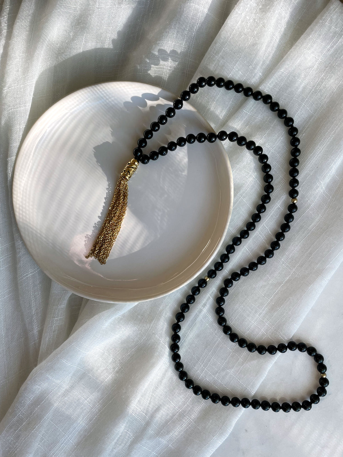 A Mala made of 6mm black Tourmaline beads with a gold plated stainless steel chain tassel on a white ceramic plate on a white light linen fabric.