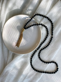 A Mala made of 6mm black Tourmaline beads with a gold plated stainless steel chain tassel on a white ceramic plate on a white light linen fabric.