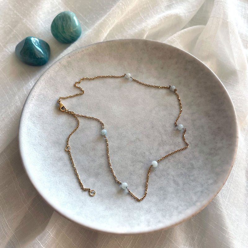 Fine golden necklace with 4mm Amazonite beads on a grey ceramic plate and two Amazonite gemstones.