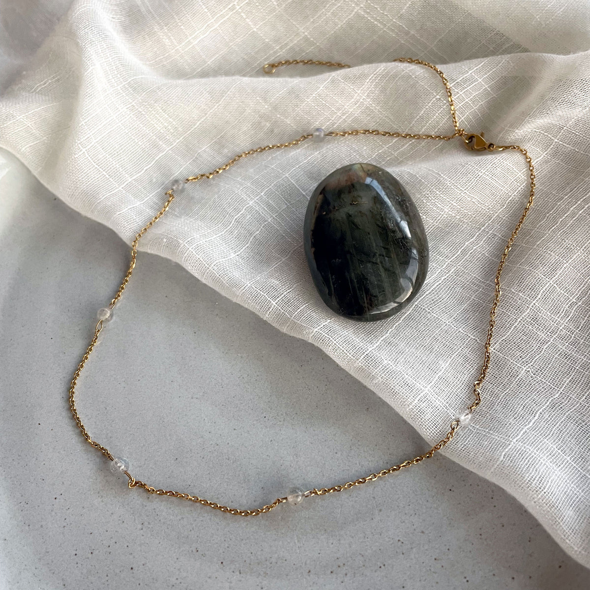A fine golden necklace with 4mm Labradorite beads on a ceramic with a white fabric with a Labradorite gemstone in the middle.