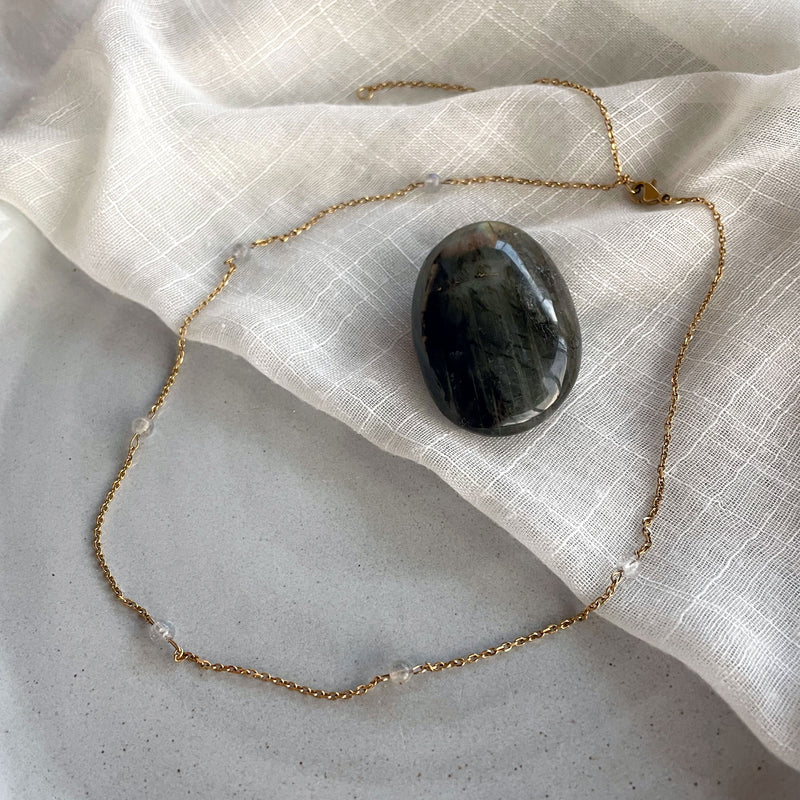 A fine golden necklace with 4mm Labradorite beads on a ceramic with a white fabric with a Labradorite gemstone in the middle.