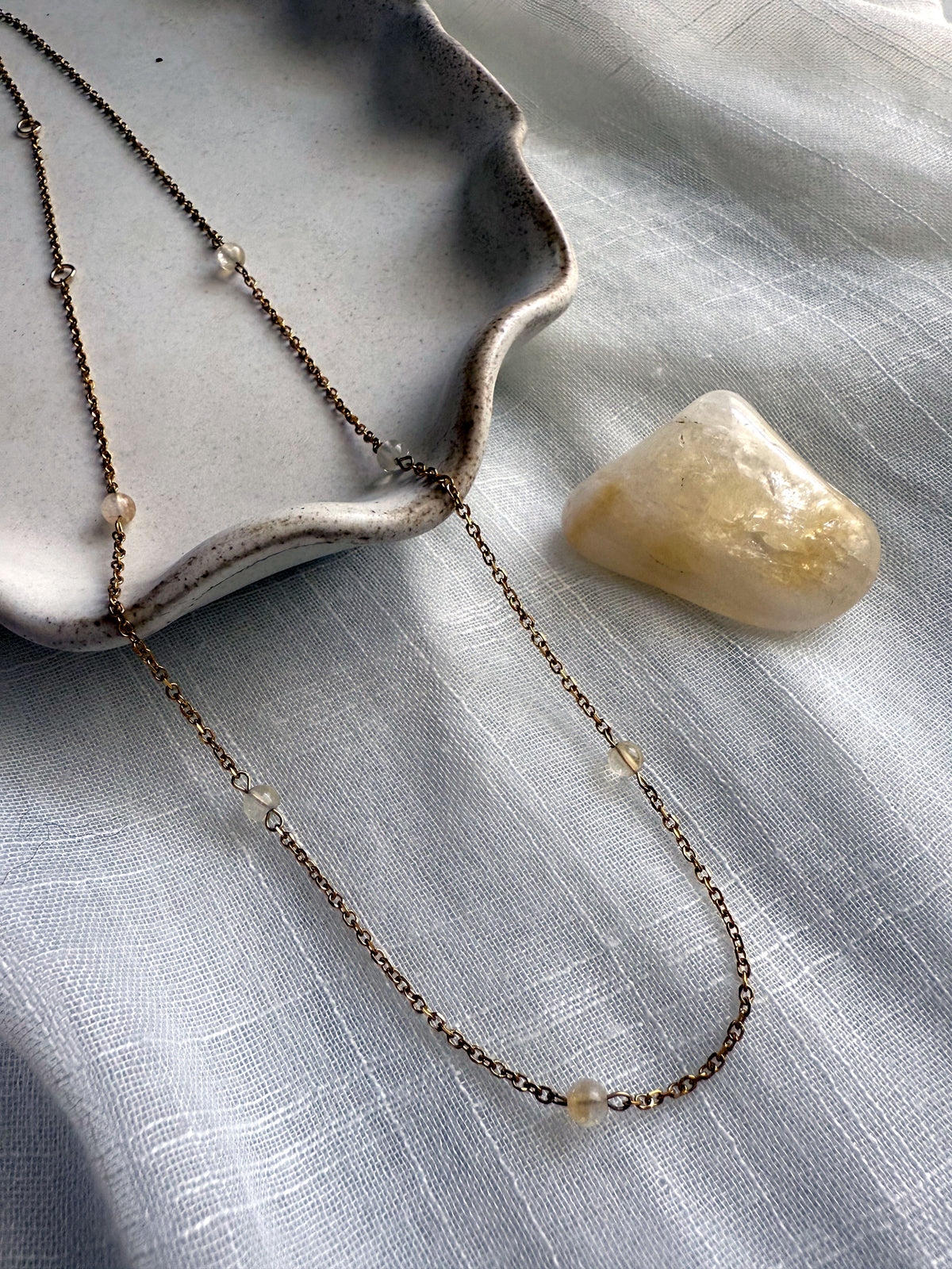 Elegant dainty necklace with fine gold chain and citrine gemstone beads, styled with a natural citrine crystal on soft fabric background.