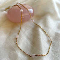 A close-up of a fine golden necklace with 6 4mm Rose Quartz beads on a light white fabric and a Rose Quartz gemstone.