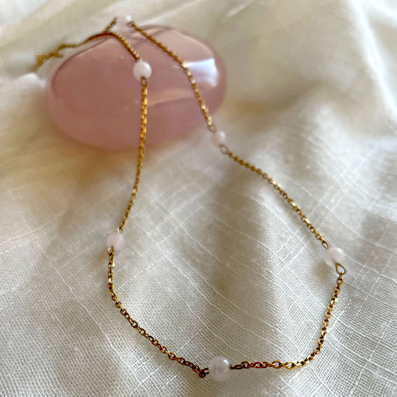 A close-up of a fine golden necklace with 6 4mm Rose Quartz beads on a light white fabric and a Rose Quartz gemstone.