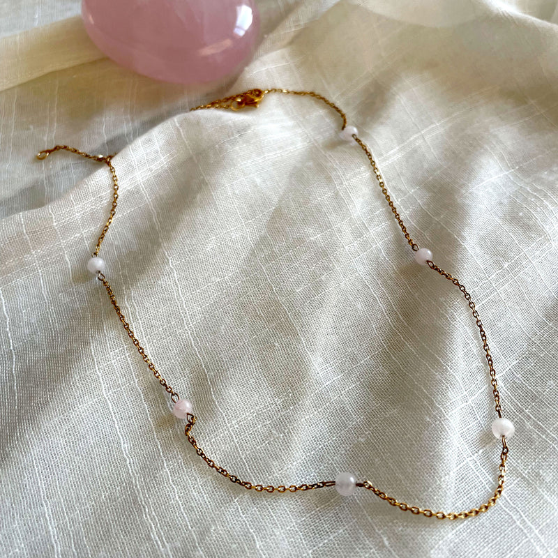 A fine golden necklace with 6 4mm Rose Quartz beads on a light white fabric and a Rose Quartz gemstone.