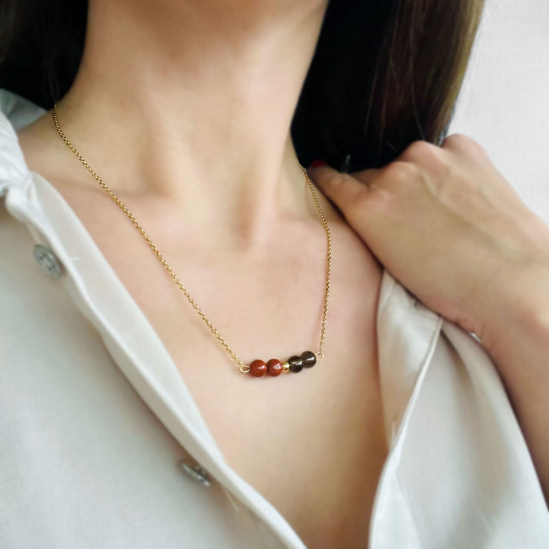 A woman wears a fine golden necklace with 2 red jasper beads, 2 smoky quartz beads and one golden bead in the middle.