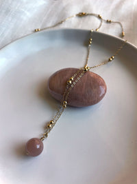 A close-up of a gold plated necklace with a 10mm peach Moonstone bead on a delicate chain with 3mm Ball and 1mm Curb Chain lying over a peach Moonstone gemstone on a white ceramic plate on a light white fabric.