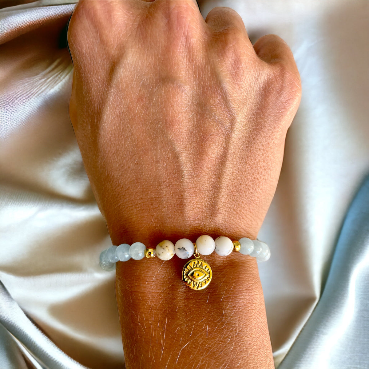 A bracelet made of Aquamarine beads, 4 white Opal beads, 2 small golden beads and a golden evil eye charm in the middle worn on a wrist.