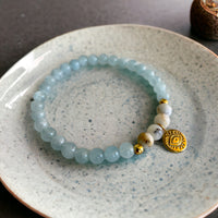 A bracelet made of Aquamarine beads, 4 white Opal beads, 2 small golden beads and a golden evil eye charm in the middle on a ceramic plate.