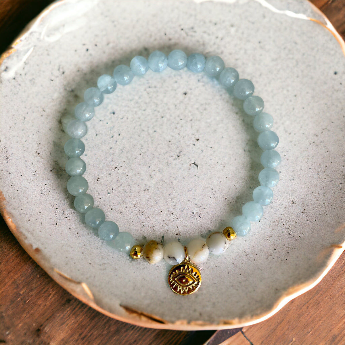 A bracelet made of Aquamarine beads, 4 white Opal beads, 2 small golden beads and a golden evil eye charm in the middle on a ceramic plate.