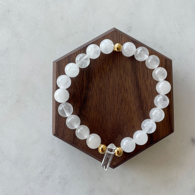 A Bracelet with 8mm Clear Quartz beads and 3 5x6mm gold plated stainless steel beads and a clear quartz tip focal point on a dark wooden display.