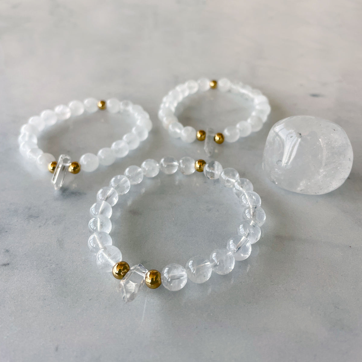 3 Bracelets with 8mm Clear Quartz beads and 3 5x6mm gold plated stainless steel beads and a clear quartz tip focal point on a marble countertop.