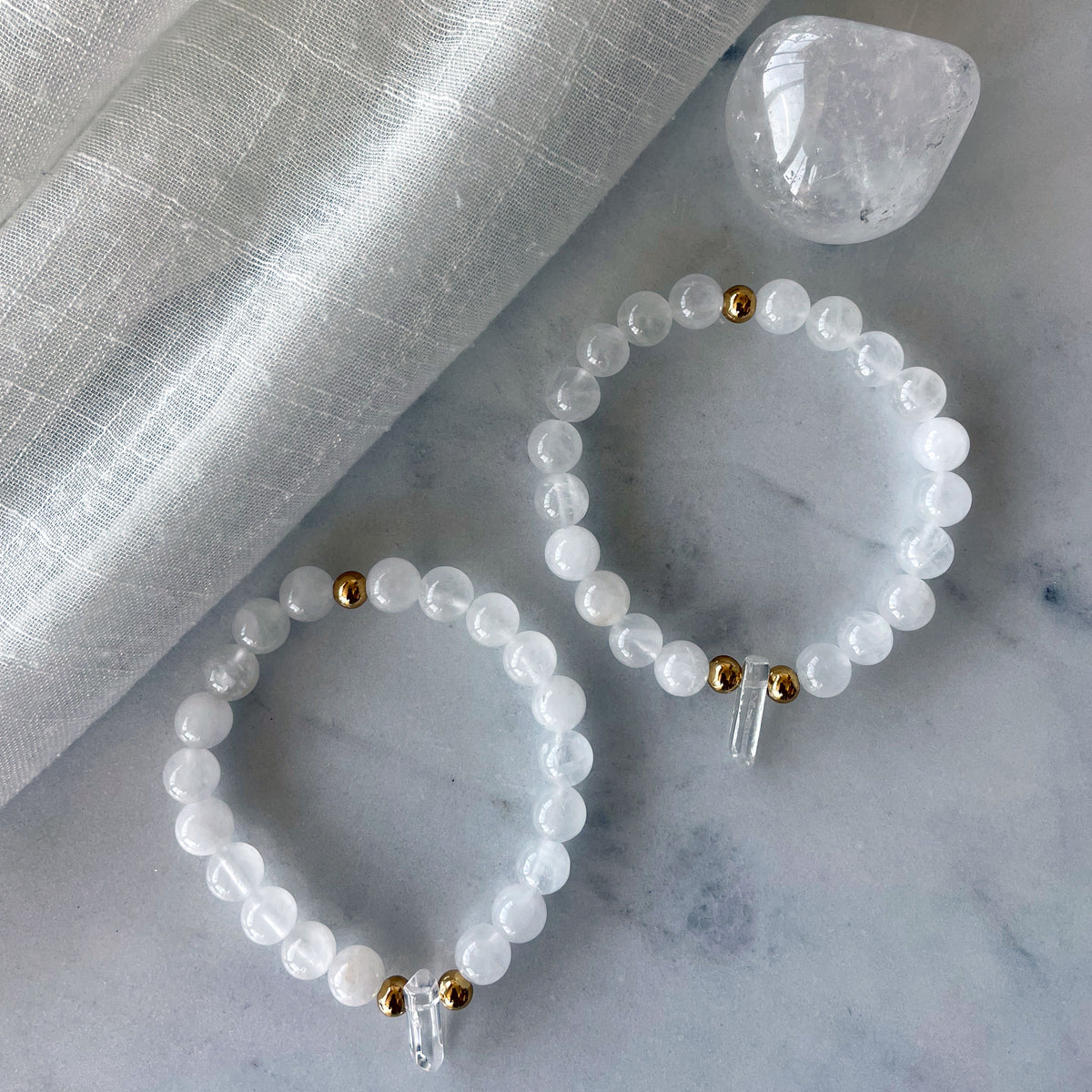 2 Bracelets with 8mm Clear Quartz beads and 3 5x6mm gold plated stainless steel beads and a clear quartz tip focal point on a marble countertop next to a clear quartz gemstone.