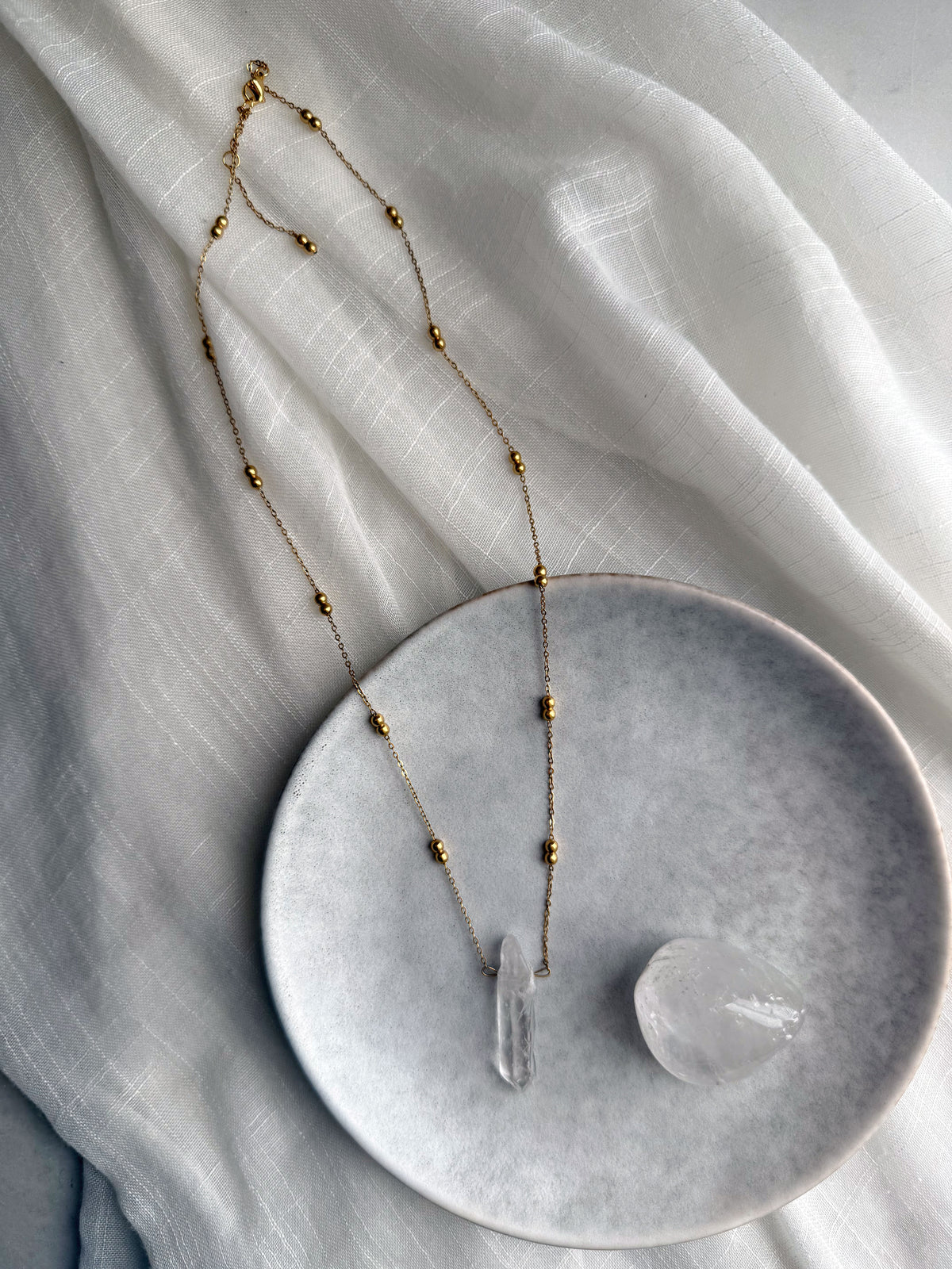 Long gold-plated beaded chain necklace with a raw clear quartz pendant, styled on a soft white fabric with a polished quartz crystal on a ceramic plate for a modern, spiritual jewelry flatlay.