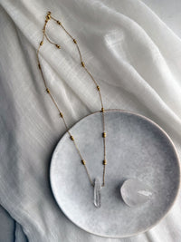 Long gold-plated beaded chain necklace with a raw clear quartz pendant, styled on a soft white fabric with a polished quartz crystal on a ceramic plate for a modern, spiritual jewelry flatlay.