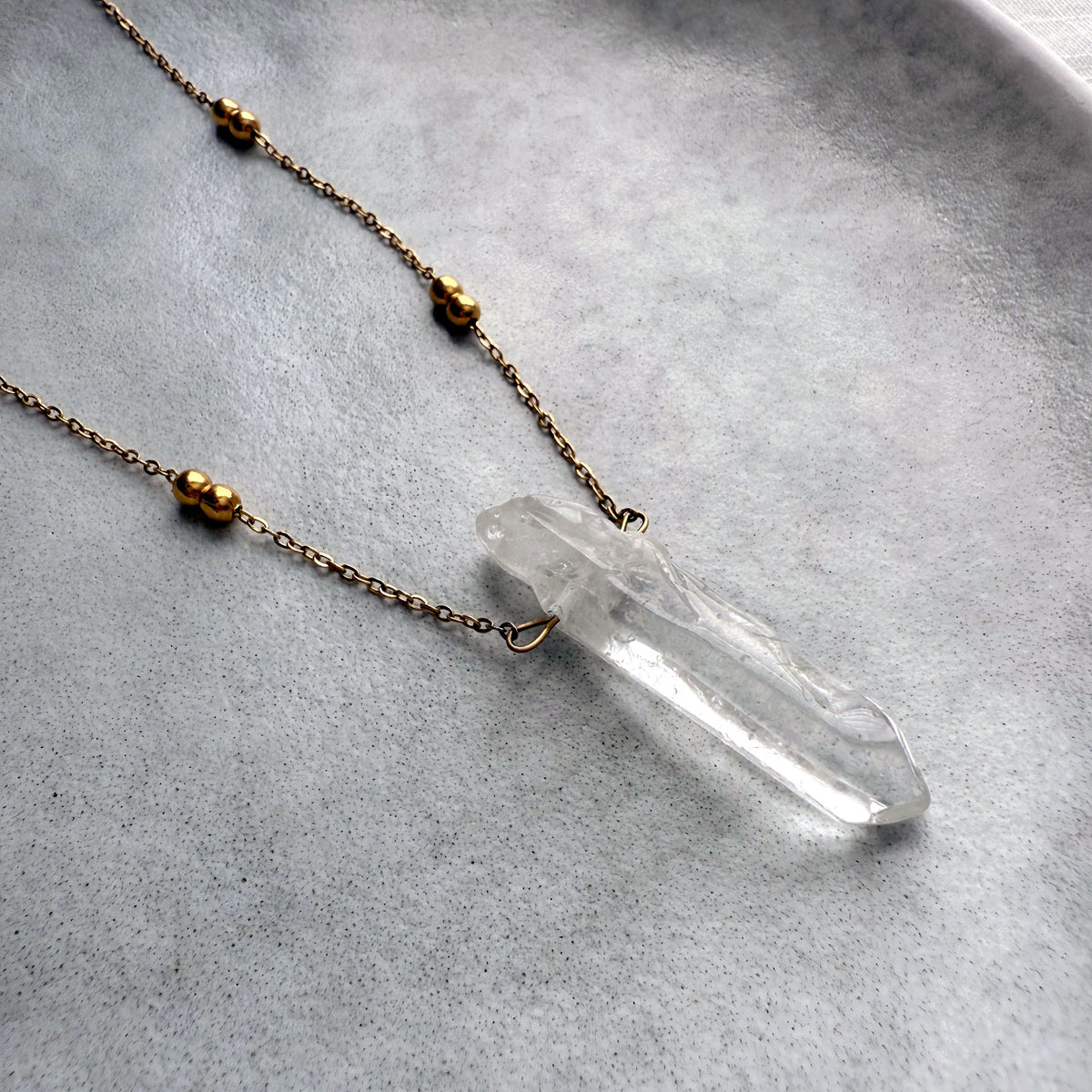 Close-up of a clear quartz crystal pendant on a delicate gold-plated chain with beaded accents, displayed on a minimalist ceramic plate for an elegant jewelry flatlay.