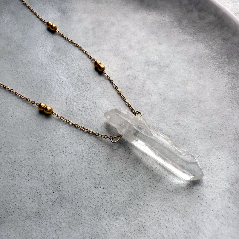 Close-up of a clear quartz crystal pendant on a delicate gold-plated chain with beaded accents, displayed on a minimalist ceramic plate for an elegant jewelry flatlay.