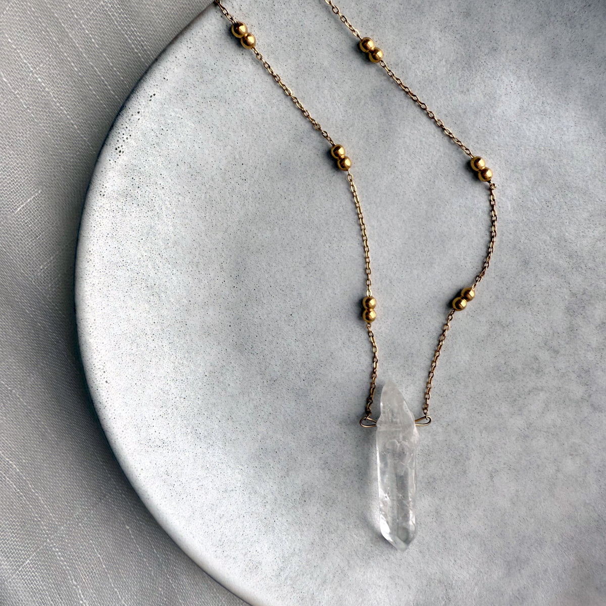 Minimalist gold-plated chain necklace with delicate round beads and a natural clear quartz crystal pendant, displayed on a textured ceramic plate for an elegant, modern jewelry showcase.