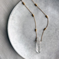 Minimalist gold-plated chain necklace with delicate round beads and a natural clear quartz crystal pendant, displayed on a textured ceramic plate for an elegant, modern jewelry showcase.
