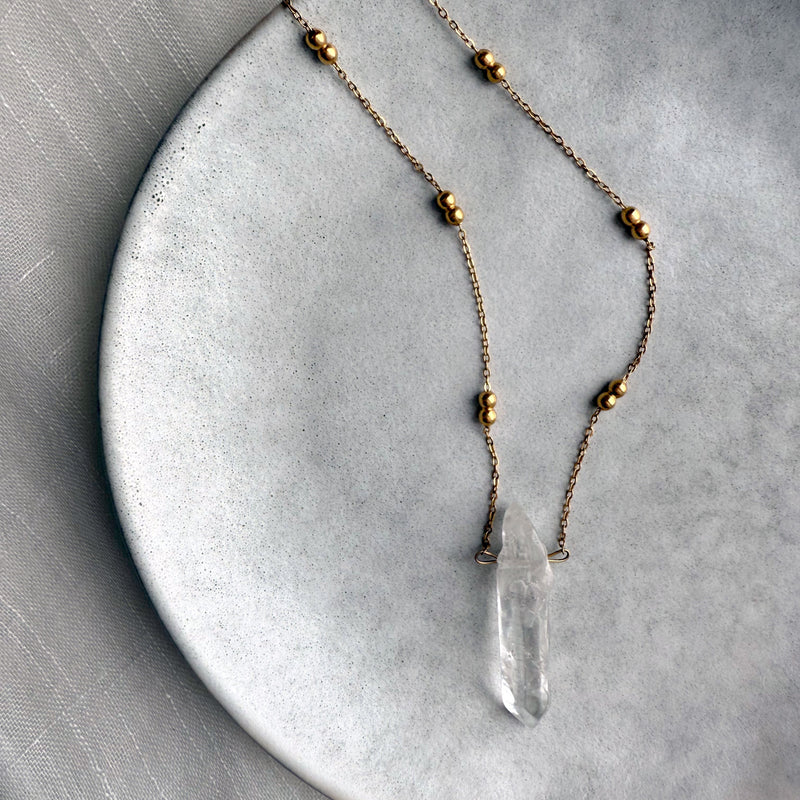 Minimalist gold-plated chain necklace with delicate round beads and a natural clear quartz crystal pendant, displayed on a textured ceramic plate for an elegant, modern jewelry showcase.