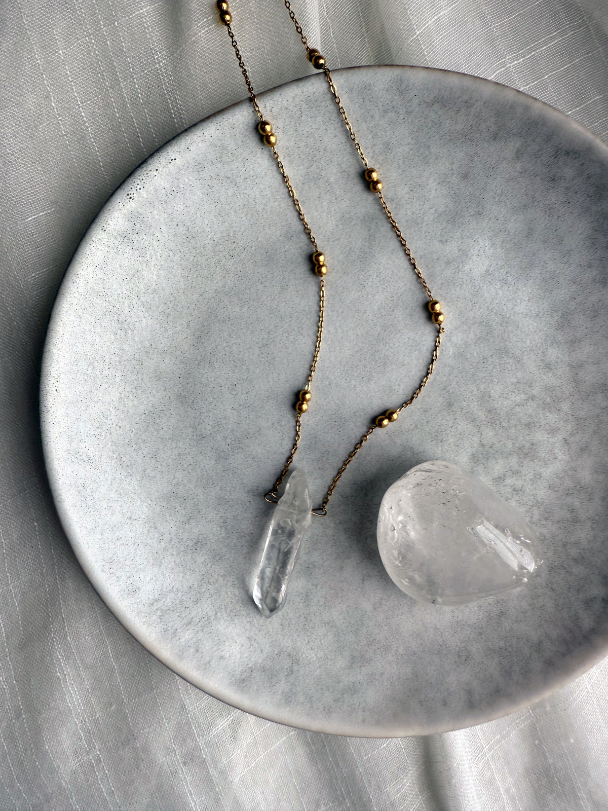 Clear quartz crystal pendant necklace on a gold-plated beaded chain, styled on a minimalist ceramic plate with a polished clear quartz stone for an elegant and spiritual jewelry display.